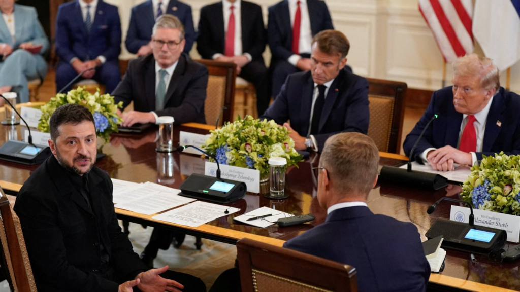 Wide shot of Volodymyr Zelensky (L) speaking to the press whilst sitting at a wooden table with sheets of paper and a mic on top. Next to him, to the right, is Finnish president Alexander Stubb. At the table, opposite them, are Keir Starmer (L), Emmanuel Macron (C) and Donald Trump