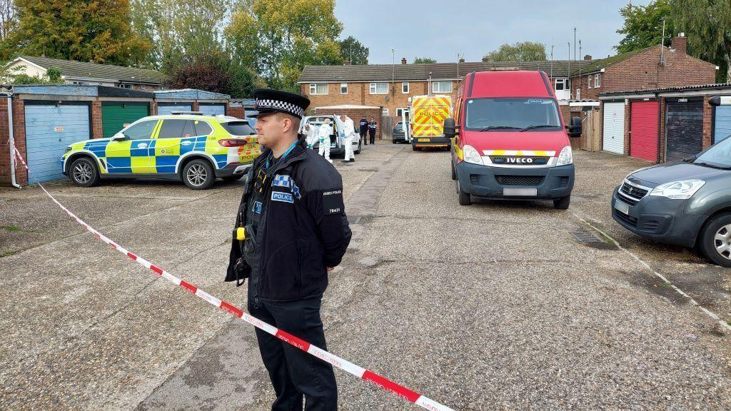 A police officer standing behind a cordon in a residential area. There is a police car and ambulance parked behind him. 