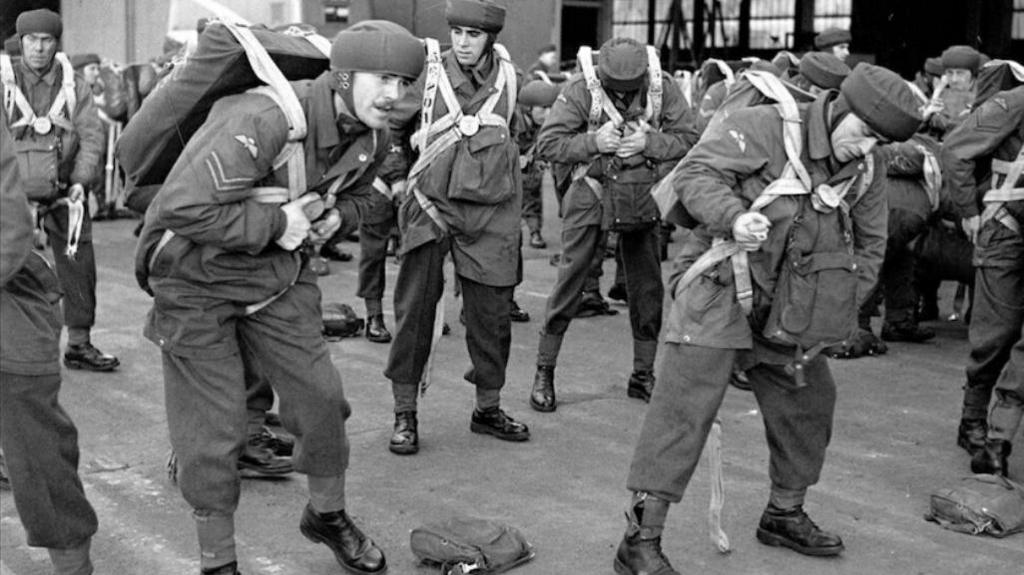 A black and white image of a group of men lined up in rows. They are wearing parachutes.