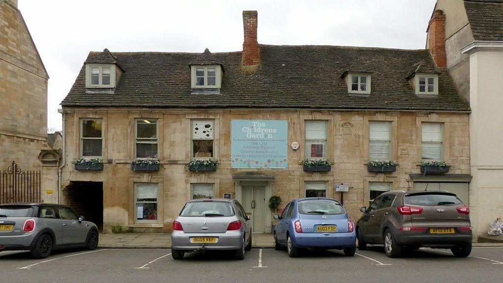 A sandstone building with a sign that reads "The Children's Garden Day Nursery". It has 4 windows in the slate roof. There are 6 windows on the first floor and 4 windows and a door on the ground floor. There is a passage through the building too. There are 4 cars parked in front in the parking spaces.
