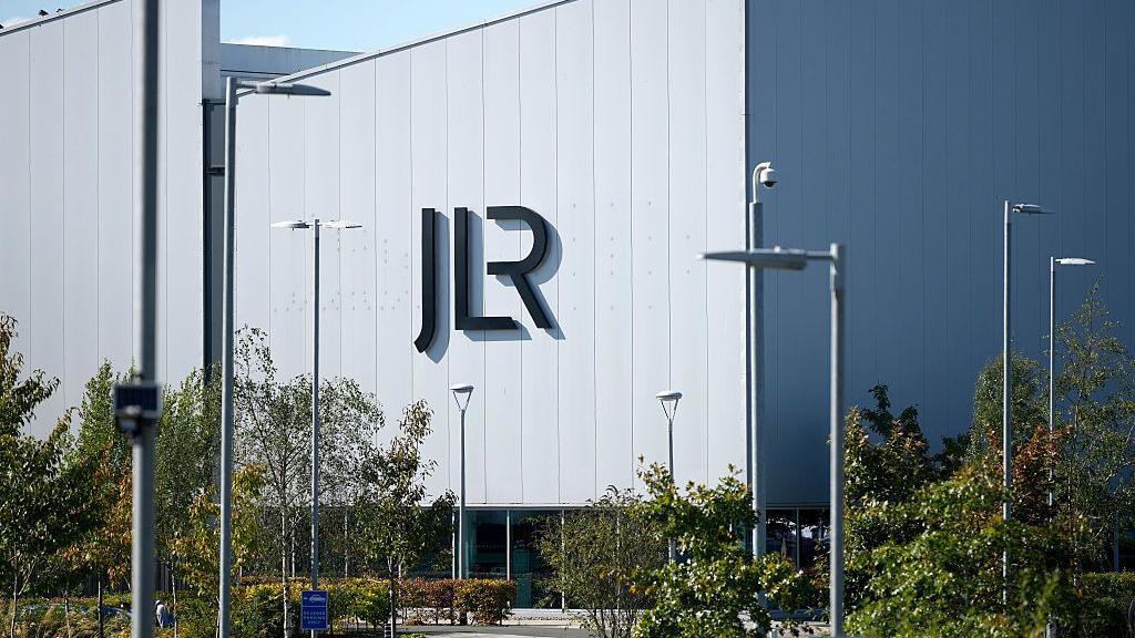 A view of a large, grey factory building sitting behind a parking lot lined with trees and shrubs. On the side of the building are large letters spelling JLR.
