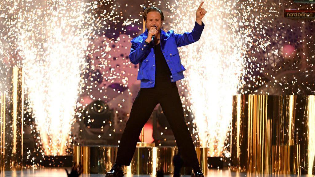 Olly Murs performs on stage during the NTAs 2024 at The O2 Arena in London. He wears a blue jacket and black trousers and he performs in front of on-stage pyrotechnics. 