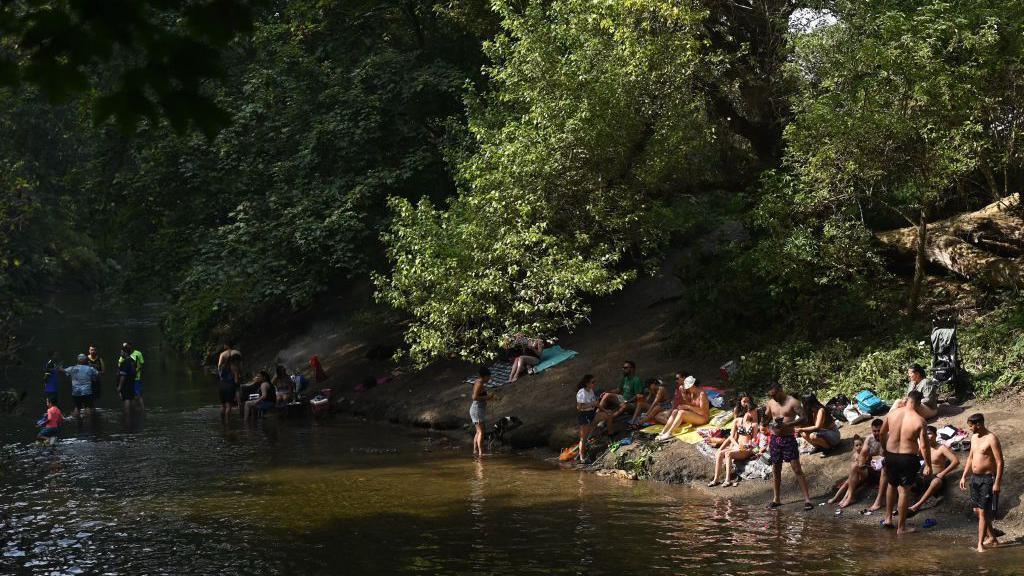 A group of people are gathered by a riverbank surrounded by lush green trees. Some individuals are wading in the water, while others are sitting or lying on towels on the sloping bank, relaxing and socializing. The scene is shaded by overhanging branches.