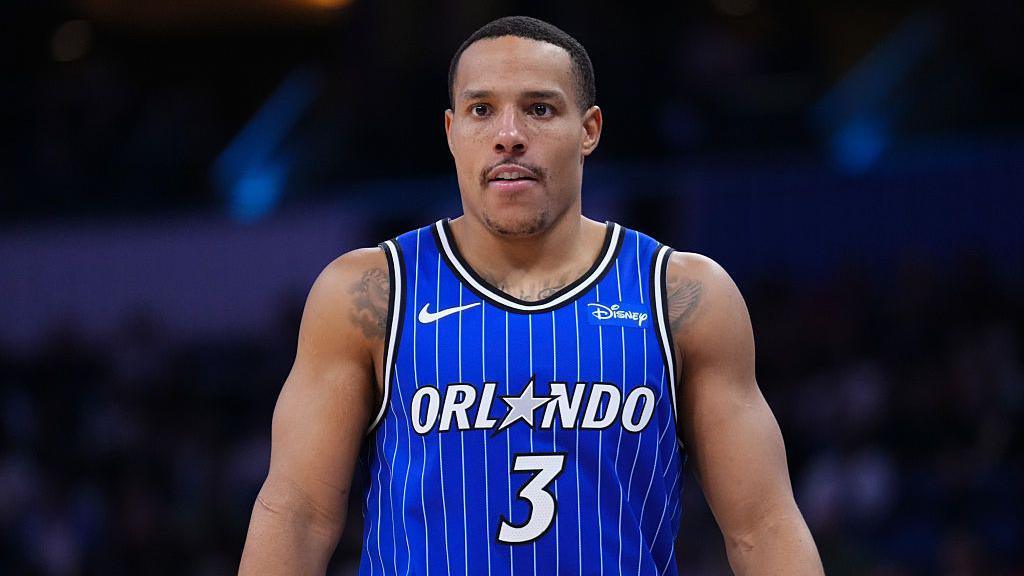 Desmond Bane seen from waist up in the centre of the image looks on in a blue Orlando Magic vest with white vertical pinstripes. A white number three is in the centre of his chest and above it the word 'Orlando' in white, with the letter A replaced by a grey five-pointed star