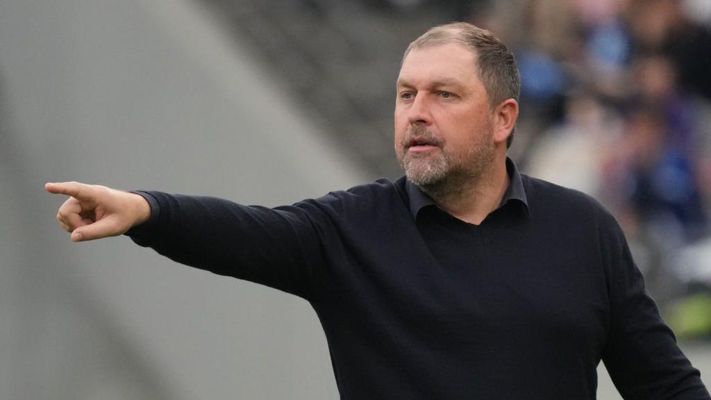 Peter Cklamovski holds his right arm out and points his finger while standing on the touchline of a match for FC Tokyo. He is wearing a black jumper with a black-collared shirt.