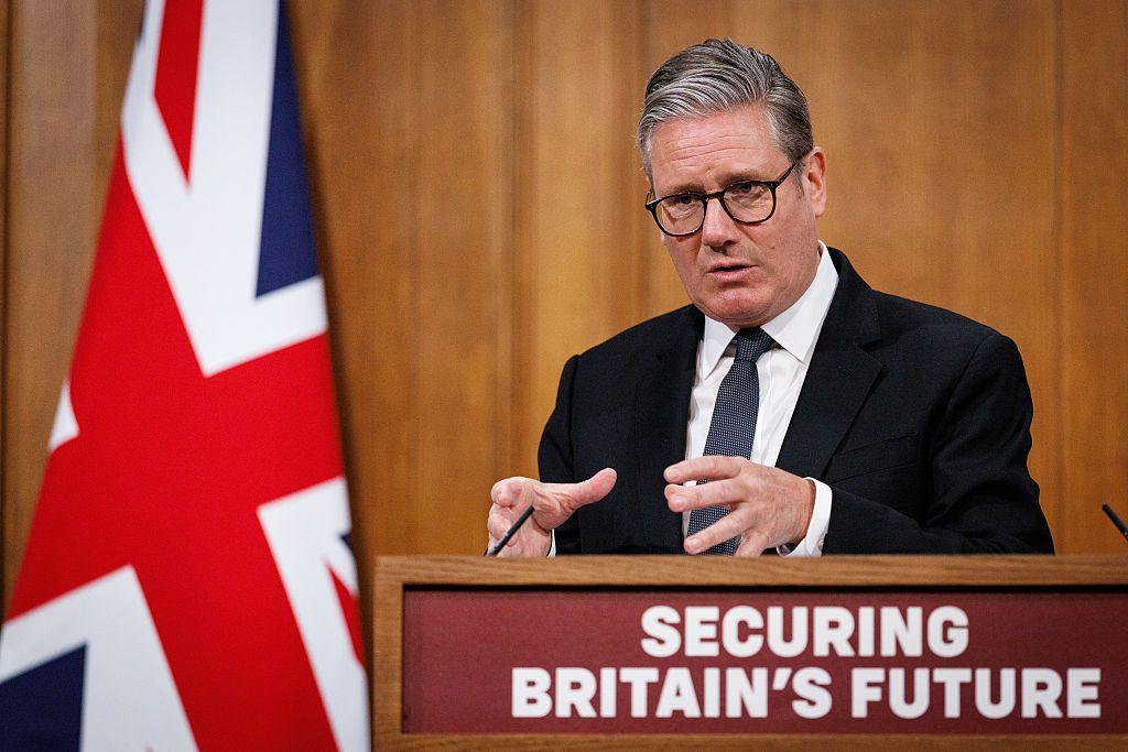 Keir Starmer is speaking behind a lectern. He has a serious, concerned expression and is using his hands to gesticulate. The lectern bears the slogan "Securing Britain's Future". Behind him, the Union Jack.