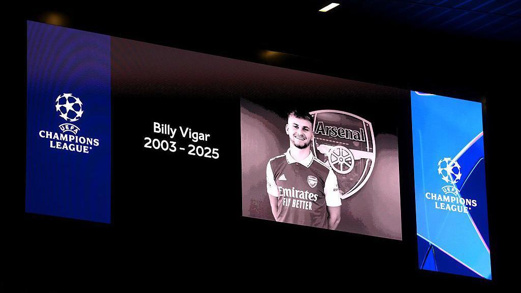 An LED screen with a greyscale image of Billy Vigar wearing an Arsenal top with white sleeves in front of the Arsenal badge, which features the word Arsenal and a canon. Next to the image is the words Billy Vigar 2003 - 2025 and two UEFA Champions League logos on blue backgrounds.