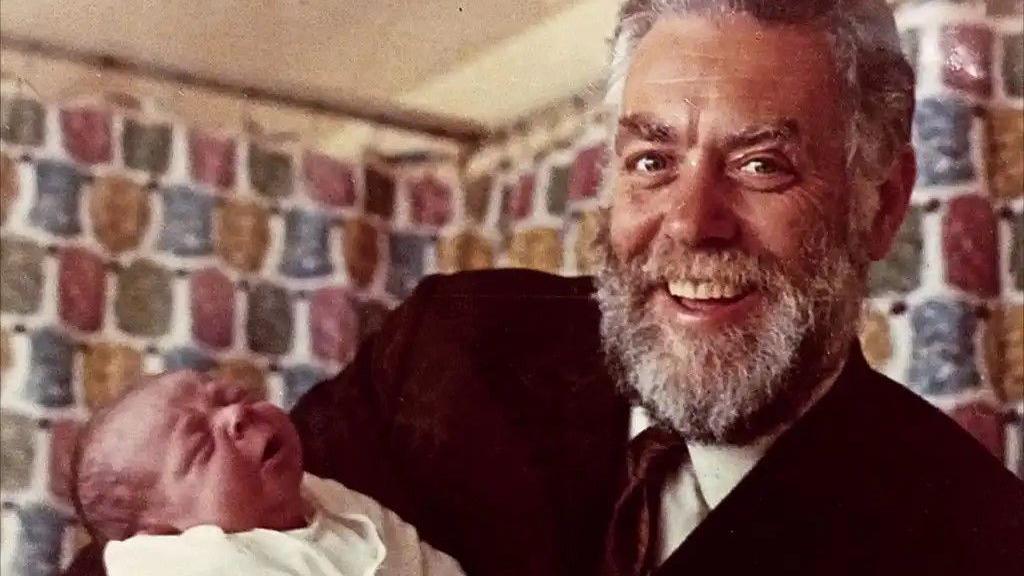 A man with grey and black beard wearing a suit smiles at the camera holding a new-born baby. The baby is crying and screwing up its face. It has a small amount of dark hair. The room behind them is colourful.