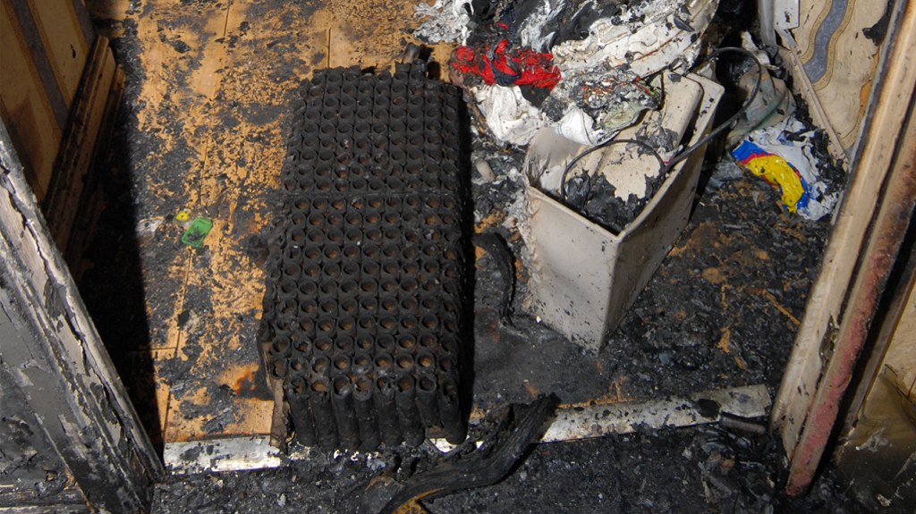 The remains of the firework show a grid of tubes. It is photographed on the floor in the house with the rubble and debris from the fire around it.