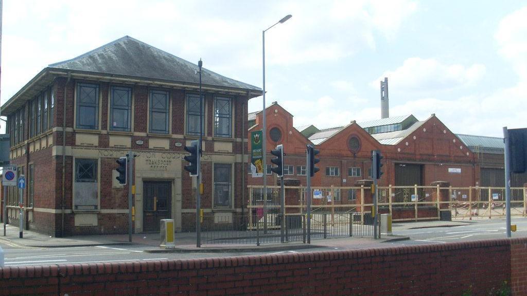 Two-storey square brick-built office building with letters above the door reading "NORTHAMPTON CORPORATION / TRANSPORT / OFFICES". The frames of the windows and the door are cream in colour.  There are triangular-roofed brick sheds to the right.  There is a road junction with traffic lights in the foreground.