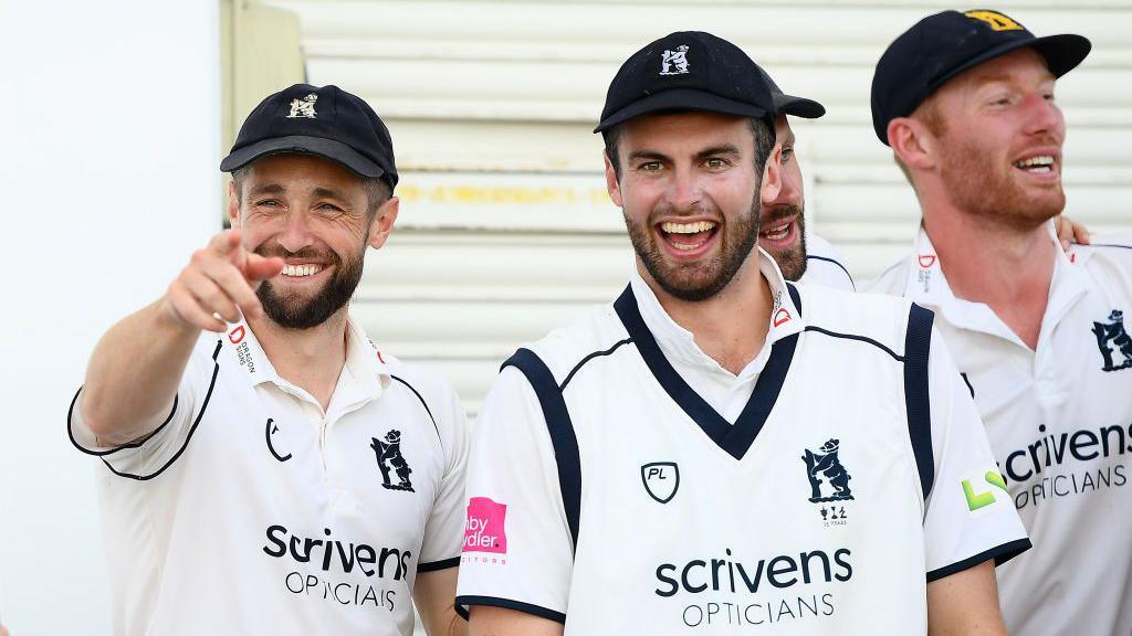 Warwickshire bowler Chris Woakes pointing at camera alongside three teammates including Dom Sibley, all wearing sleevless cricket whites and blue club caps 