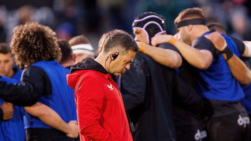 Johann van Grann walking with his head down on the pitch with the Bath players in a huddle with arms around each other's shoulders in the background behind him