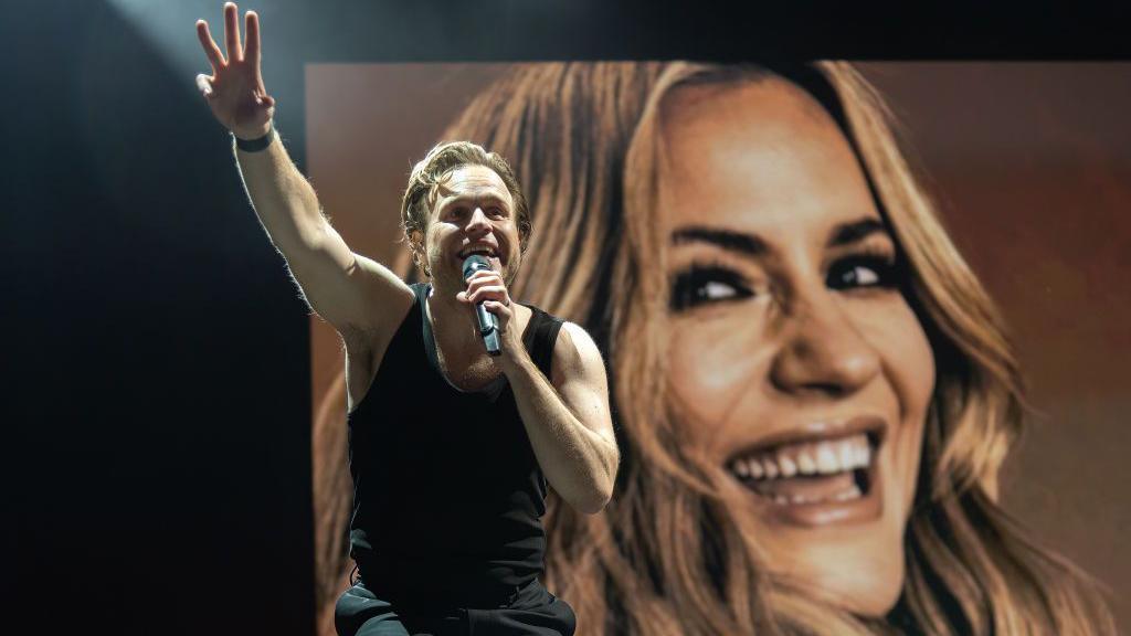 Olly Murs, singing into a microphone and in front of a picture of Caroline Flack, performs at Flackstock 2024 at Englefield House on July 22, 2024