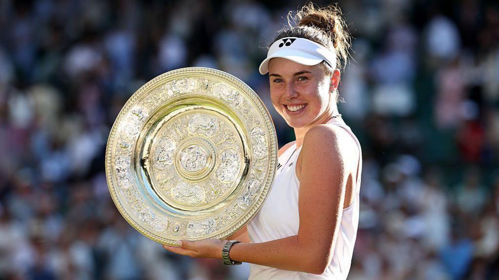 Linda Noskova of Czechia poses for a photo with the Venus Rosewater Dish. She is smiling holding the trophy. She is wearing a white tennis dress, a white cap and a silver watch. It is a big circle shaped trophy that is gold and silver.
