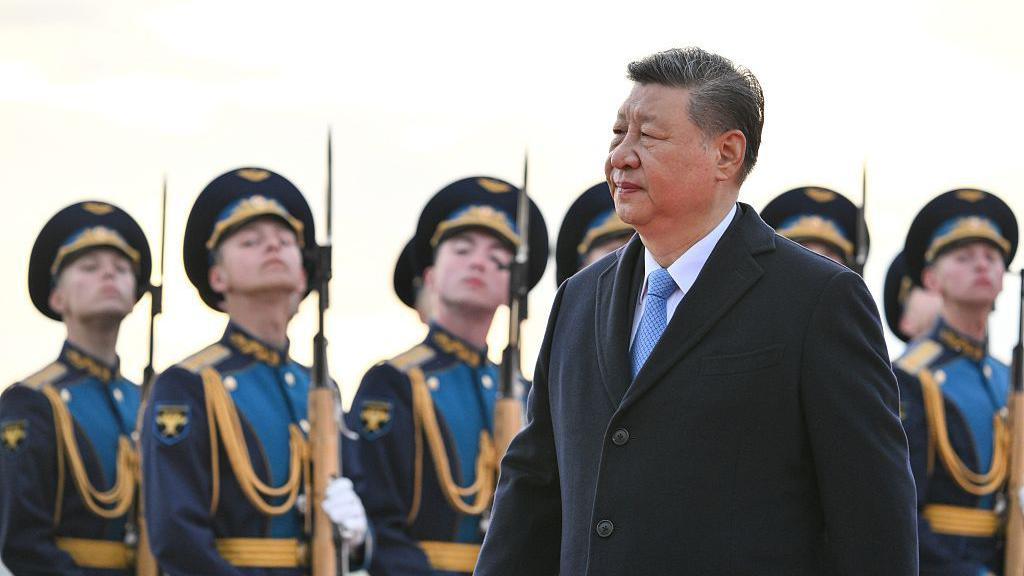 Xi Jinping wearing a black coat and light blue tie, walking in front of a row of Russian soldiers carrying bayonets
