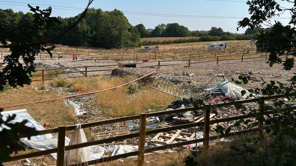 Abandoned land with hard standings and green/yellow grass growing. There is an abandoned rotating-style clothes line and rubbish in the foreground, and a caravan in the mid-distance. A lot of wooden three-bar fencing has been erected. There are green trees in the background.