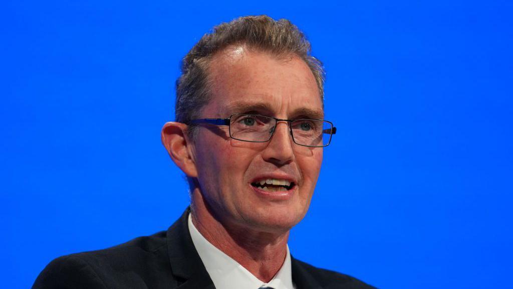 David TC Davies standing in front of a blue background. He has short grey hair, rectangle glasses and is wearing a suit