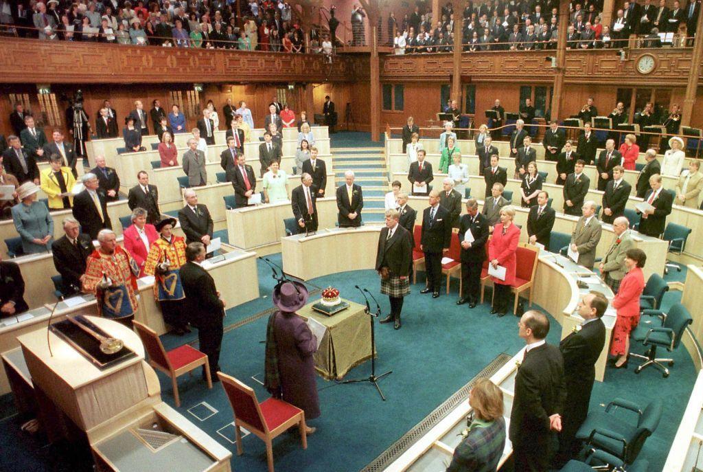 Queen Elizabeth, wearing a purple hat and coat, stands in the centre of the parliament chamber with her back to the camera.  A crown is on a table in front of her. Around the chamber, politicians and the Royal Party are standing.