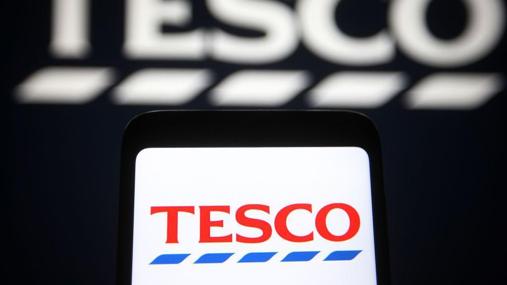 The Tesco logo is displayed on a smartphone, against a black background displaying a larger, blurred version of the supermarket's logo.