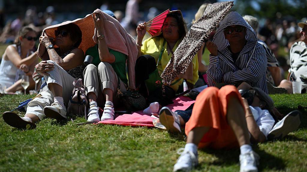 Spectators seen hiding from the sun by placing jackets over their heads and holding up umbrellas as they sit on the grass