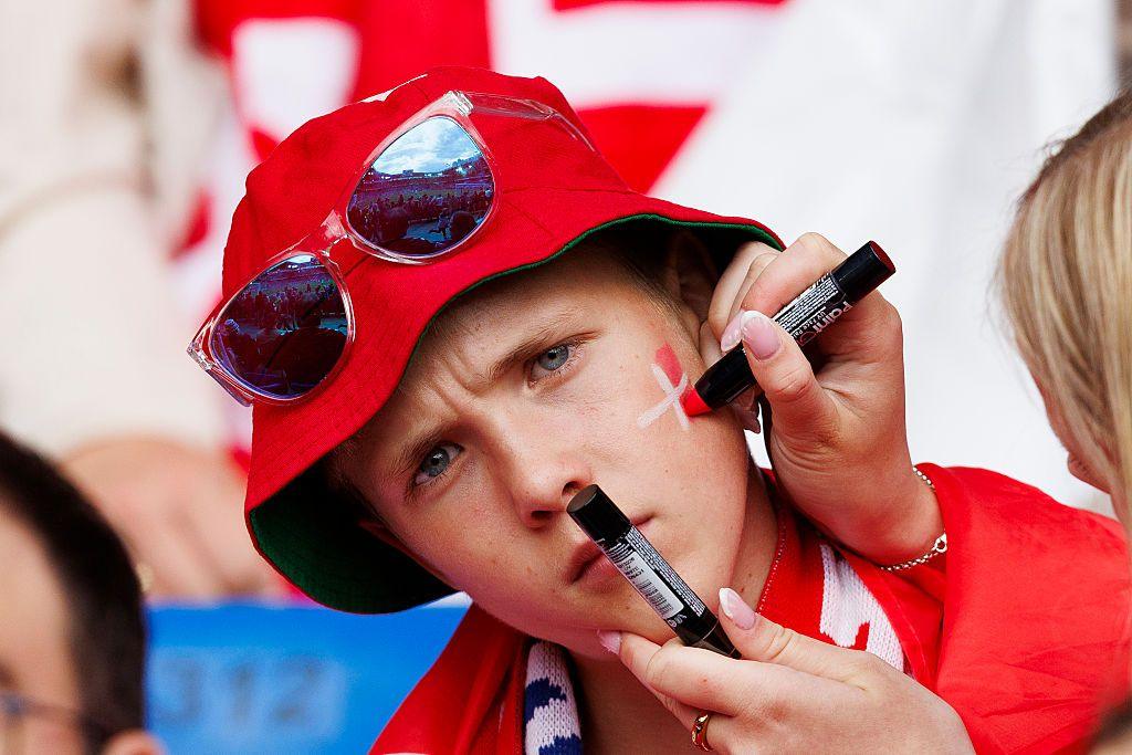 A woman paints the Danish flag on a young man's cheek. He is wearing a Danish scarf, a bucket hat and sunglasses