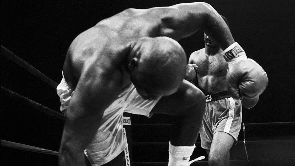 Joe Frazier falls into the camera with George Foreman in the background