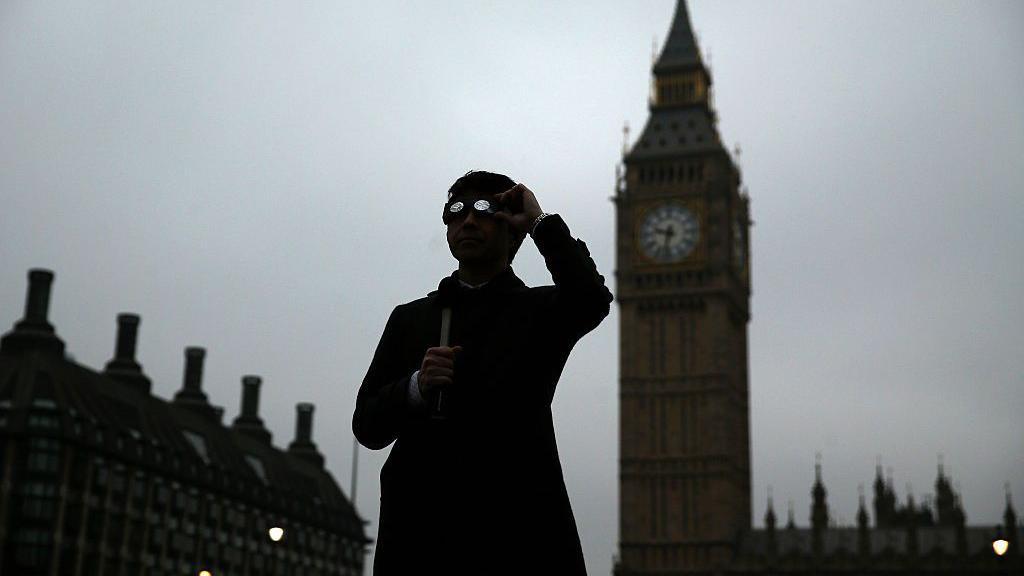 Man wearing eclipse-viewing glasses stands in front of the Elizabeth Tower and the Palace of Westminster in central London.