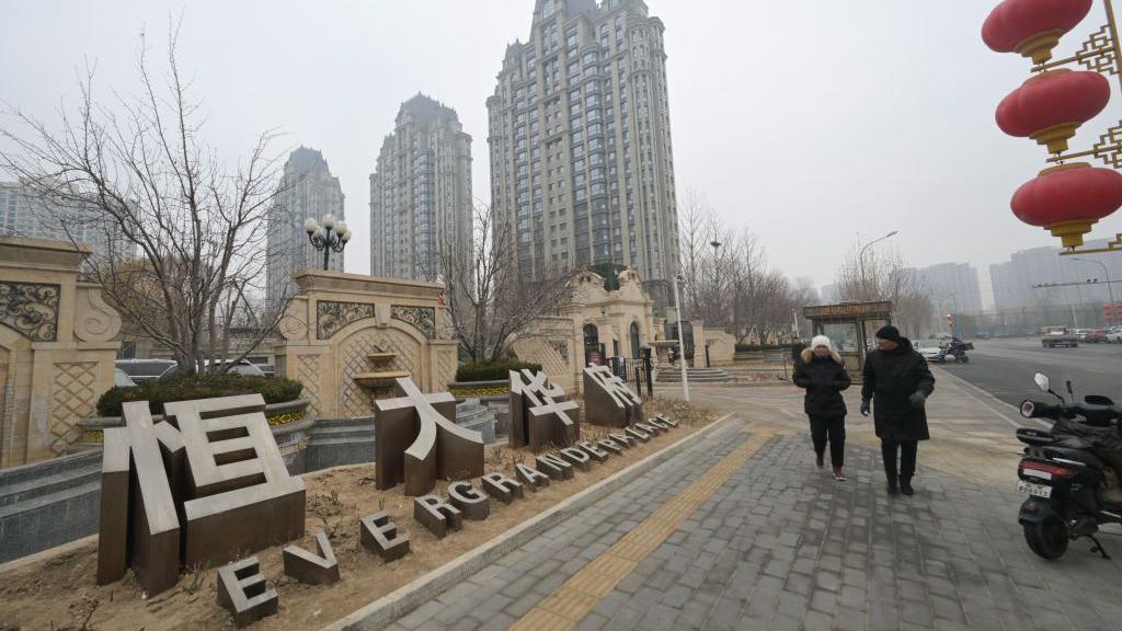 People wearing coats and hats walk past an Evergrande Group residential complex called Evergrande Palace in Beijing on 30 January, 2024 on a misty overcast day.