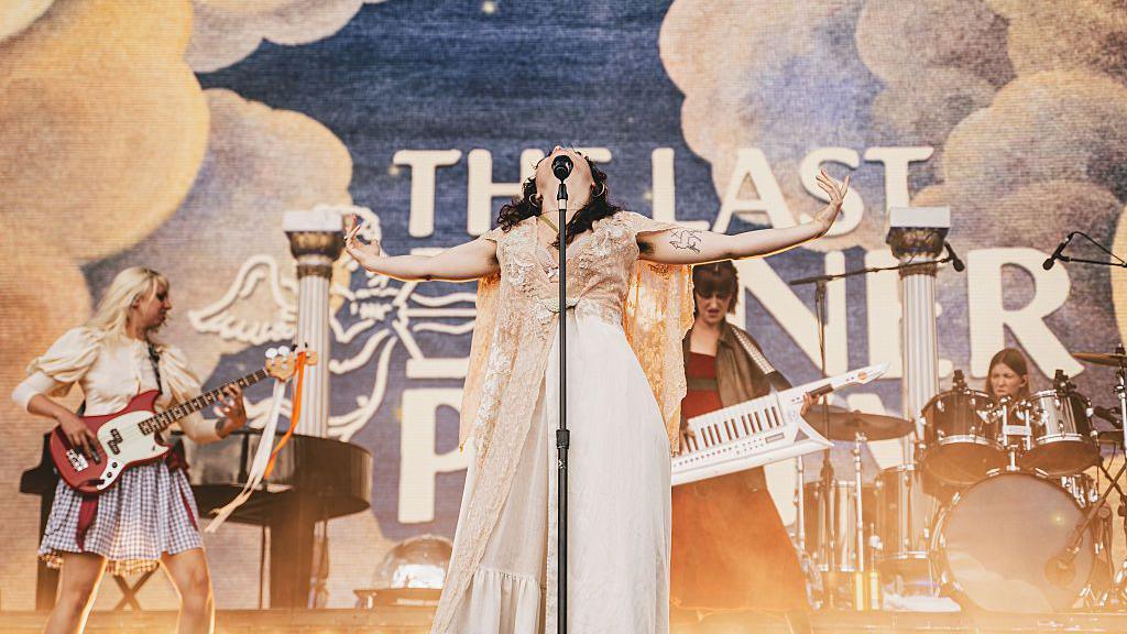 The Last Dinner Party performs onstage during Tramlines at Hillsborough Park. The lead singer outstretches her arms and throws her head back.
