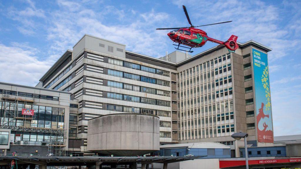 University Hospital Wales, as seen from the Accident & Emergency entrance. The image shows a ten-storey building, with a lower section to the left and a higher, L-shaped section to the right, both in grey brick and glass. A red rescue helicopter is hovering above the entrance area, poised to land.