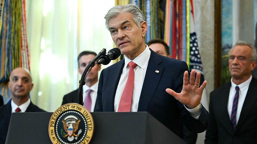 Medicare and Medicaid Administrator Mehmet Oz speaks in the Oval Office during an event about weight-loss drugs at the White House in Washington, DC on November 6, 2025. He is speaking at a podium that features the seal of the President of the United States. His hair is short and grey, and he is wearing a black suit with a red tie.