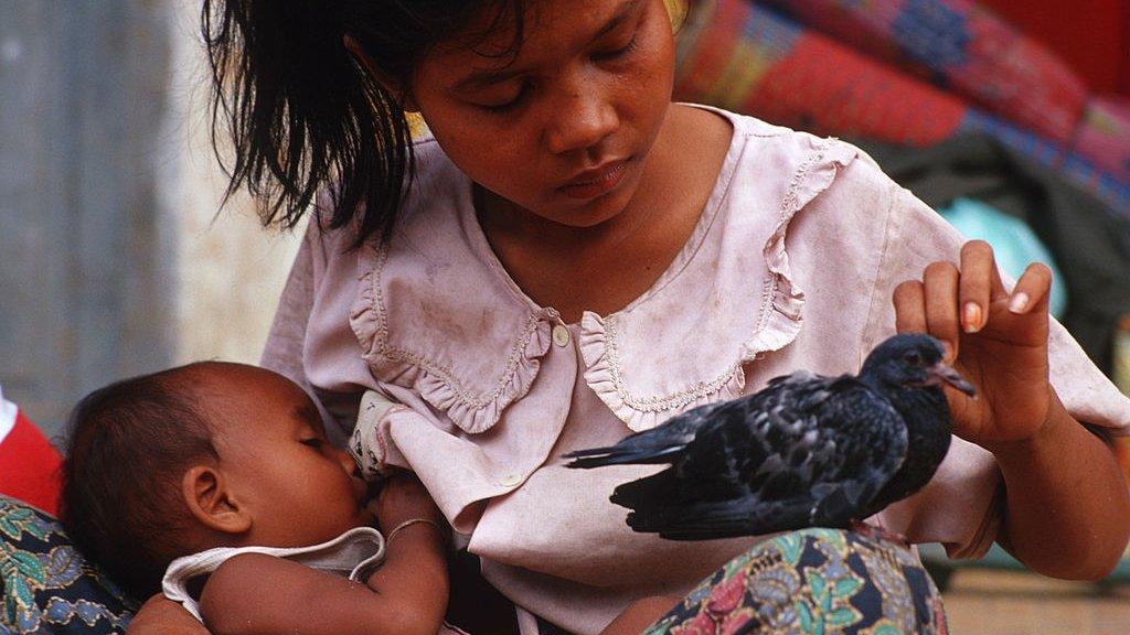 A young Cambodian mother feeding her baby and petting a pigeon