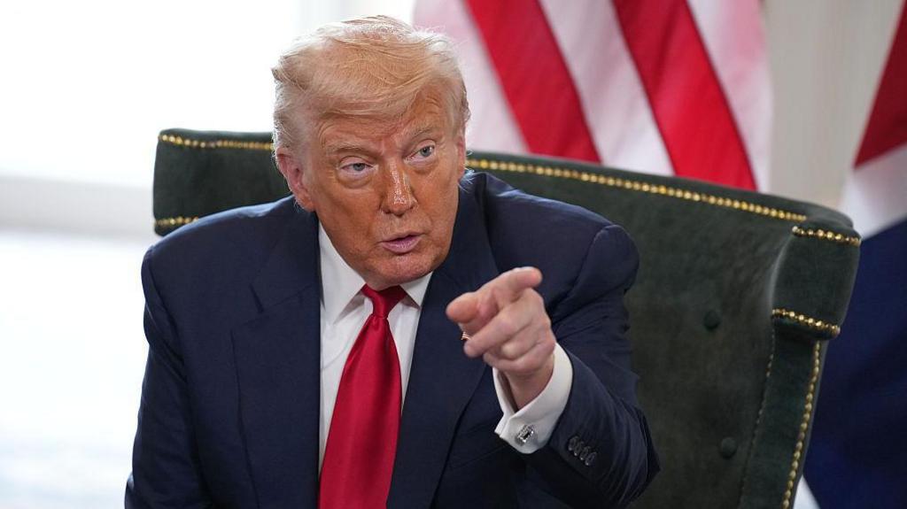 Trump, wearing a blue suit jacket, red tie and white shirt, pointing at something off-camera while sat in the ballroom of his Turnbury golfing resort in Scotland