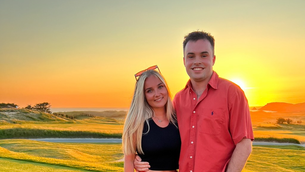 Jack and Emily holding each other and smiling for the camera. Jack is wearing a pink button down t shirt and has one arm around Emily's waist. He has short cropped brown hair. Emily is wearing a fitted black sleeveless top. She has straight blonde hair down below her shoulder and has sunglasses on the top of her head. They are standing on groomed grass in front of a golf bunker with sun setting in the background