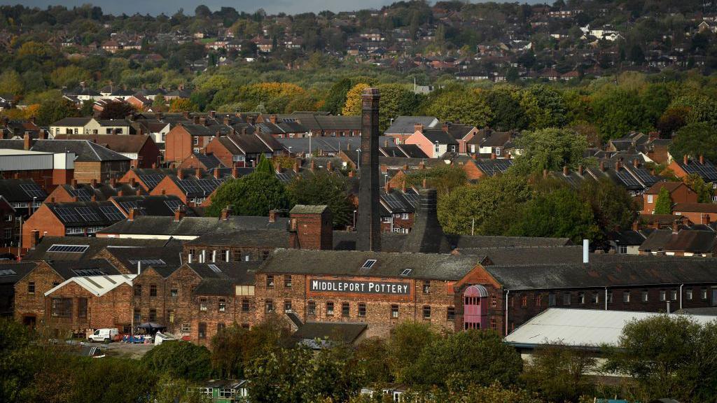 Middleport Pottery seen from a distance, surrounded by other industrial buildings with houses and trees behind it extending up a hill. The brown brick building has a large Middleport Pottery sign in black and white high up. A chimney and kiln can be seen behind it.