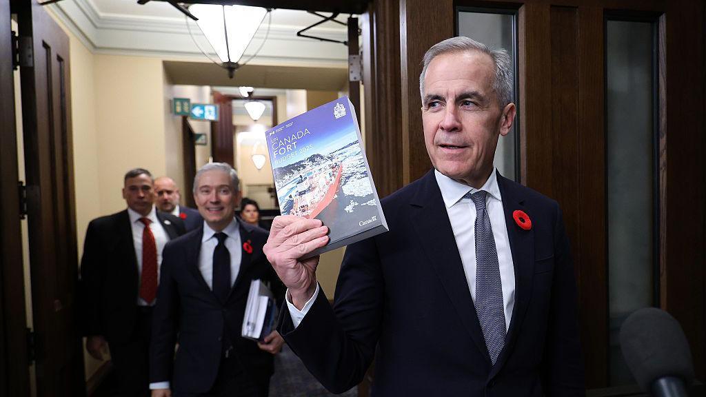 Mark Carney, Canada's prime minister,in a dark suit, holds up a copy of the federal budget. Walking behind him are members of his cabinet, including his finance minister.
