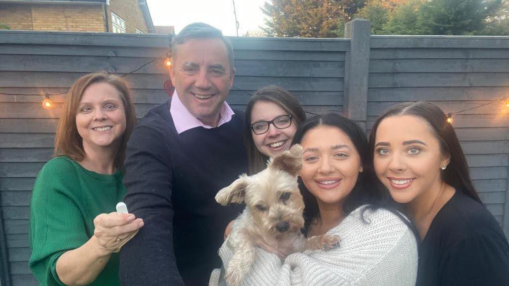A photo of the Hunt family in a garden with their small, Yorkshire Terrier-type dog. Carol, left, wears a green top, John in the centre wears a pink shirt and navy blue jumper, Amy in the centre wears glasses, Louise to the right wears a white jumper and is holding the dog, and Hannah, far-right, is wearing a navy blue top. All five family members are smiling warmly.