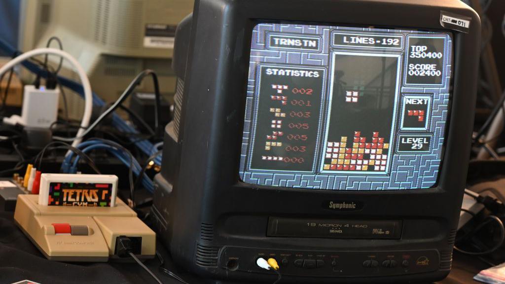 An NES console with a Tetris cartridge inserted sits on a table next to an old Symphonic TV showing Tetris being played on screen.