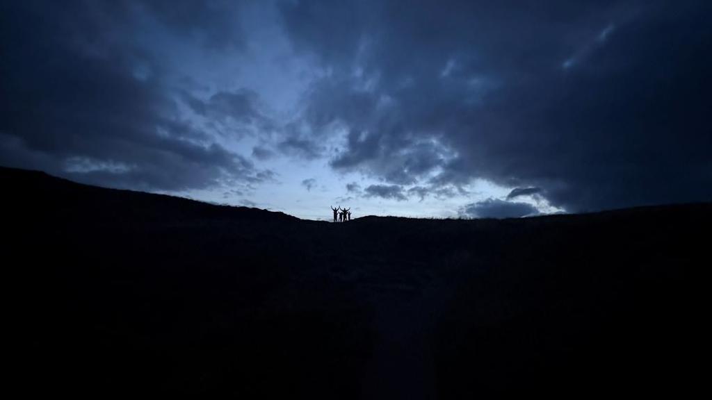 Four silhouetted people in the far distance wave at the camera from a hill