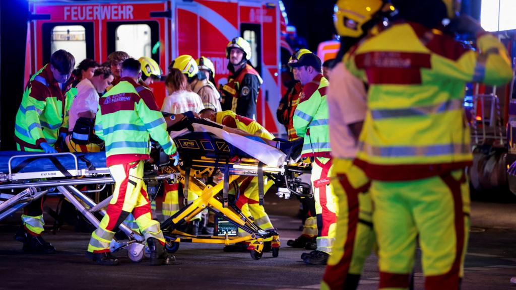A number of emergency services personnel standing near an ambulance, with a person on a stretcher
