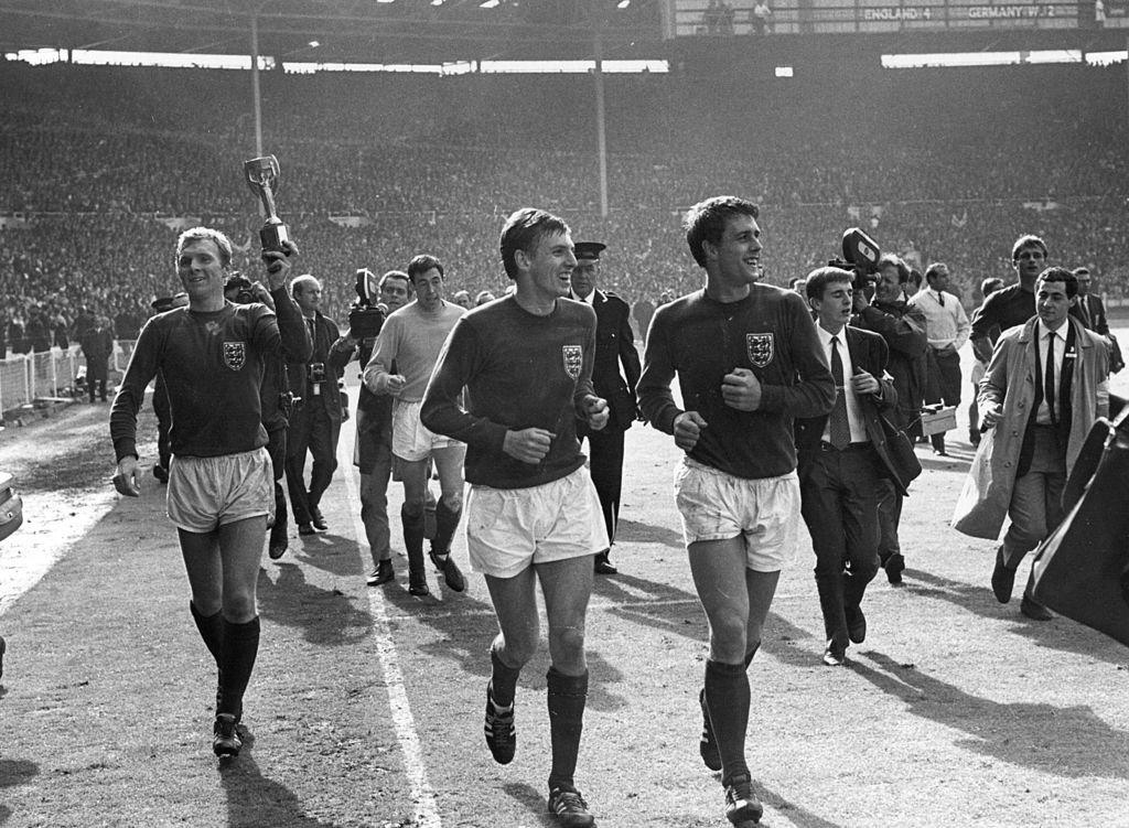 England captain Bobby Moore (1941 - 1993) (left) and colleague Geoff Hurst (right), lead the team around the pitch at Wembley, after their 4-2 victory over West Germany in extra time in the World Cup final