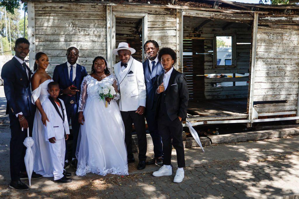 A man and woman stand next to their wedding guests under the shade of a tree and next to a wooden cabin.