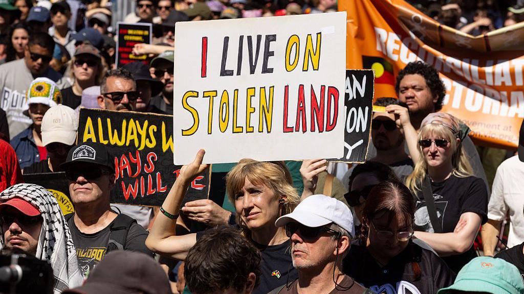 A woman in a crowd holding a sign that reads "I live on stolen land".