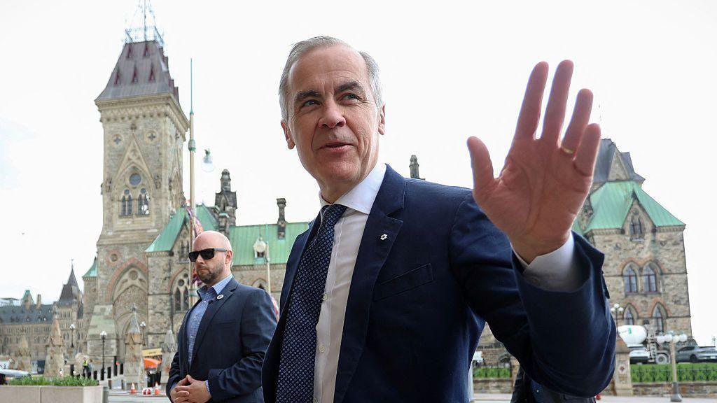 Canada's Prime Minister Mark Carney speaks to the media upon arriving at his office on Parliament Hill April 29. He is smiling and waving, and wearing a dark blue suit and white shirt. 