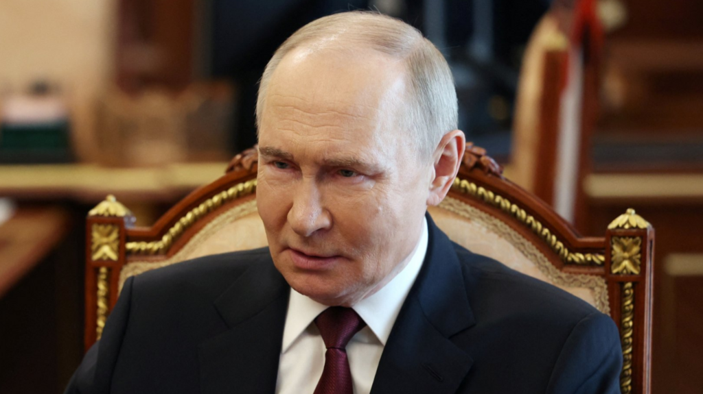 Vladimir Putin sitting down in a wooden chair in dark suit, white shirt and burgundy tie 