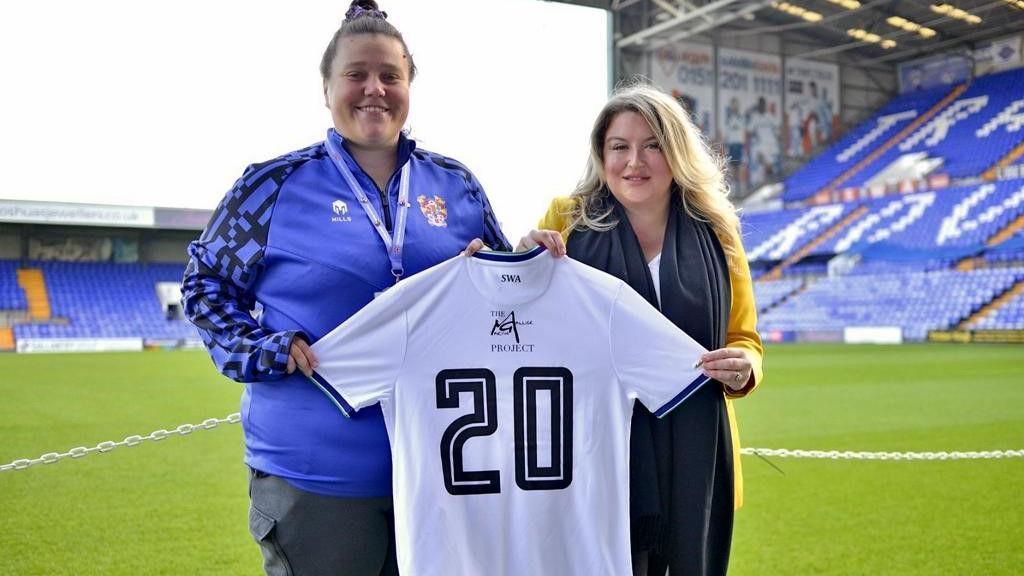 Hannah Naylor wears a Tranmere top and dark trousers, while Jessica Gallier Booth stands next to her at the Tranmere ground. Ms Gallier Booth is wearing a mustard-coloured jacket, white top and black scarf. They are holding a Tranmere Rovers women's team shirt with the number 20 and The Martin Gallier Project logo on the back. 