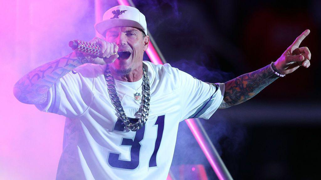 Vanilla Ice sings into a microphone He wears a white NFL jersey with the number 31 on the front, a white baseball cap and a big silver neck chain.