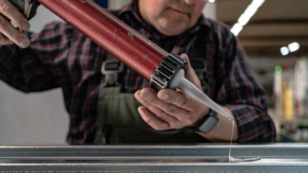 A worker squeezing a grey-coloured silicone sealant on to a metal bar.