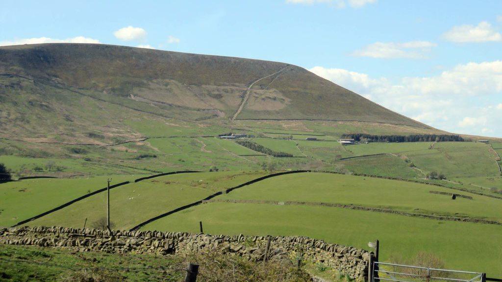 View of the green Pendle Hill on a sunny day under a blue sky. Low green fields can be seen in the foreground with a stone wall at the bottom image.