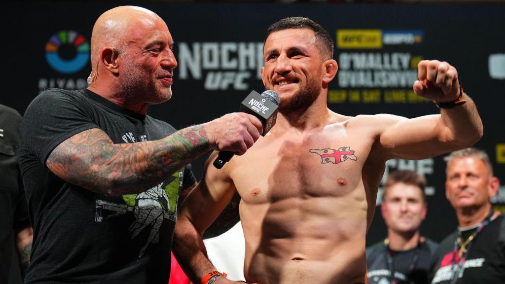 Shirtless MMA fighter Merab Dvalishvili is interviewed by Joe Rogan, wearing a black T-shirt and holding a microphone, at UFC 306 in Las Vegas, Nevada in September